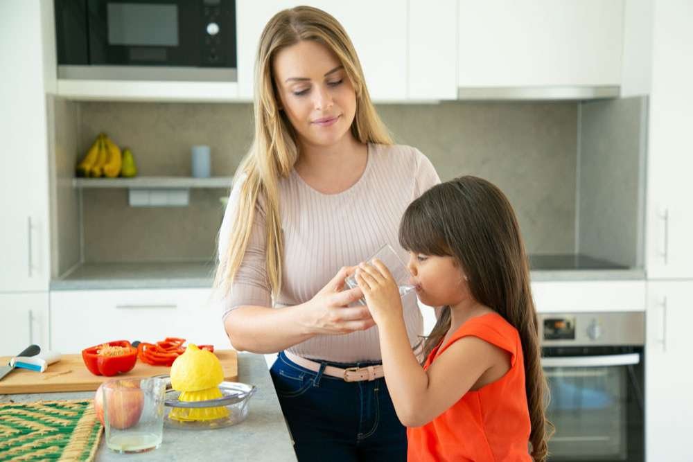 mom-giving-daughter-glass-of-water-to-drink-while-cooking-salad-and-squeezing-lemon-in-kitchen-family-cooking-or-healthy-lifestyle-concept